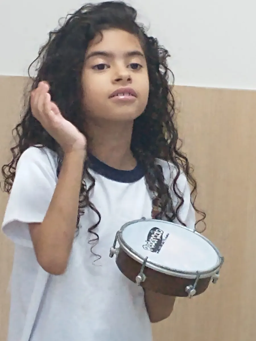 Menina segurando um pandeiro com a mão junto ao ouvido, atenta ao som