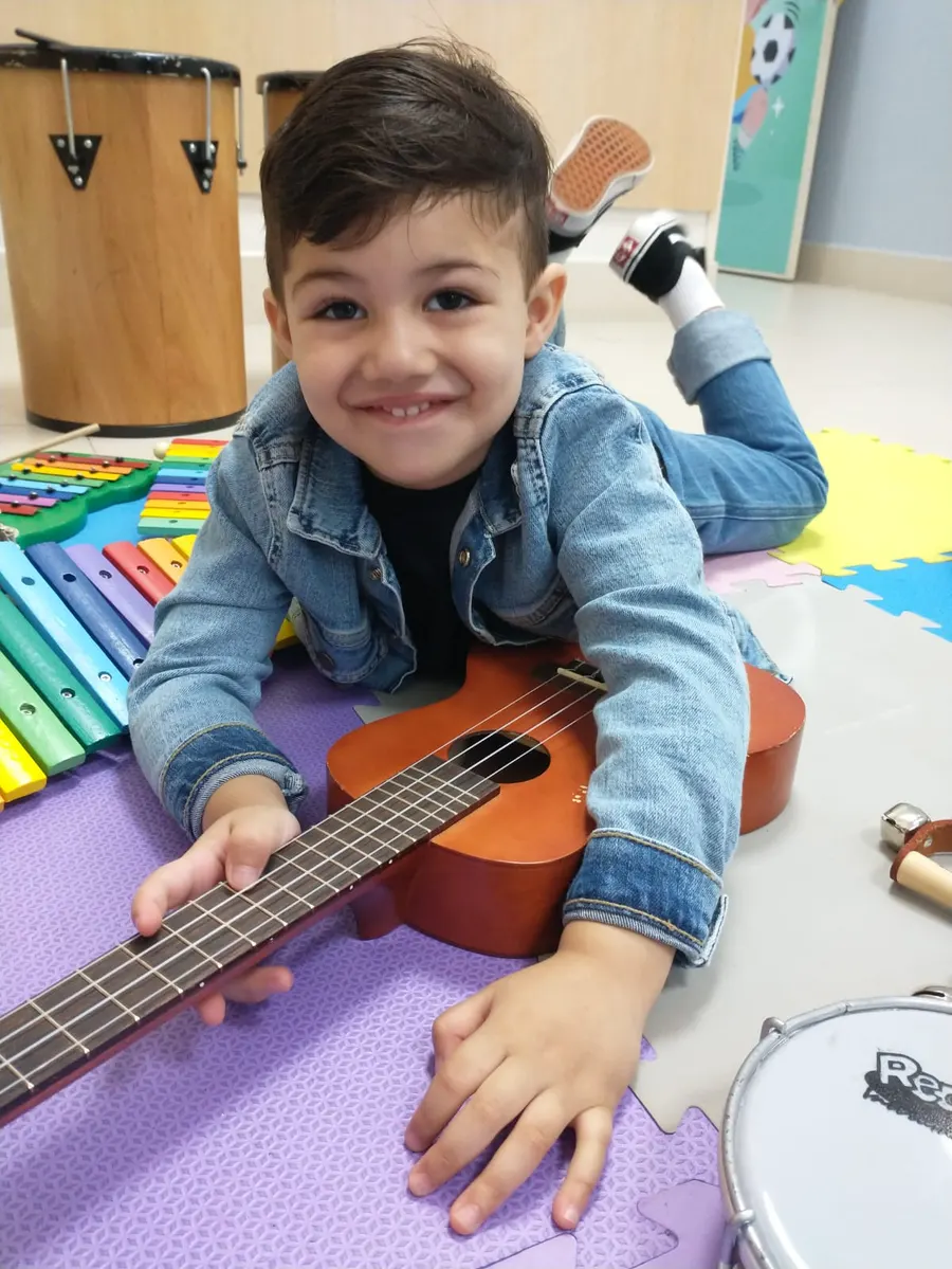 Menino deitado no tapete tocando um ukulele, ao lado de um xilofone colorido